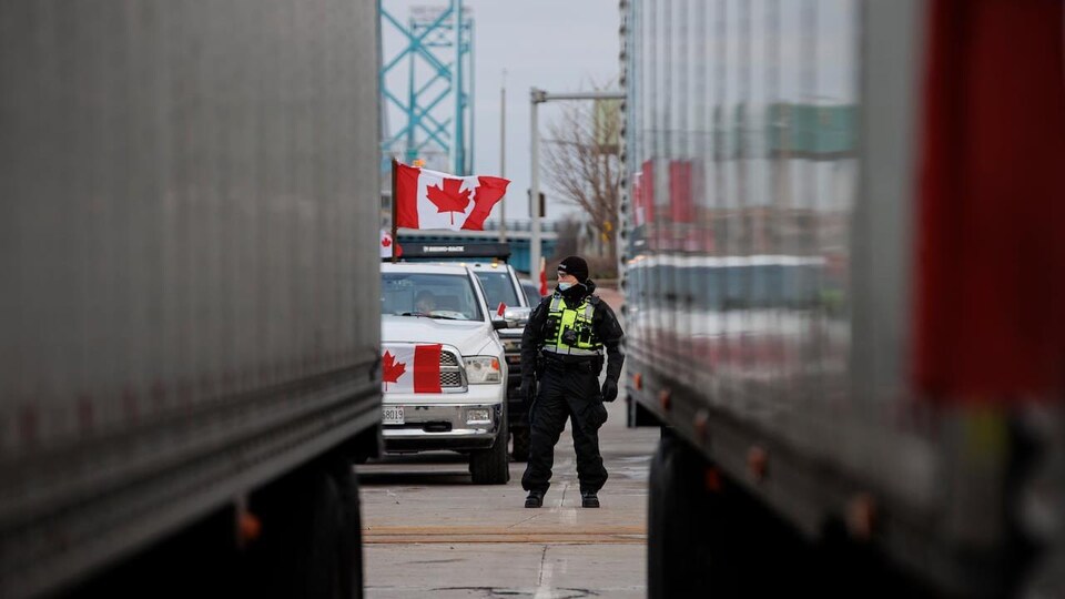 Un policier est sur la voie qui mène à l'entrée du pont Ambassador. Derrière lui, des véhicules arborent des drapeaux canadiens. Le photographe est installé entre deux camions.