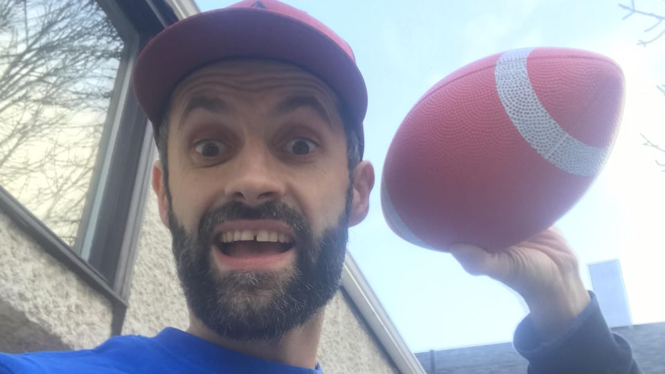 Un homme vêtu d’un chandail bleu et d’une casquette rouge qui tient dans sa main gauche une balle de football.