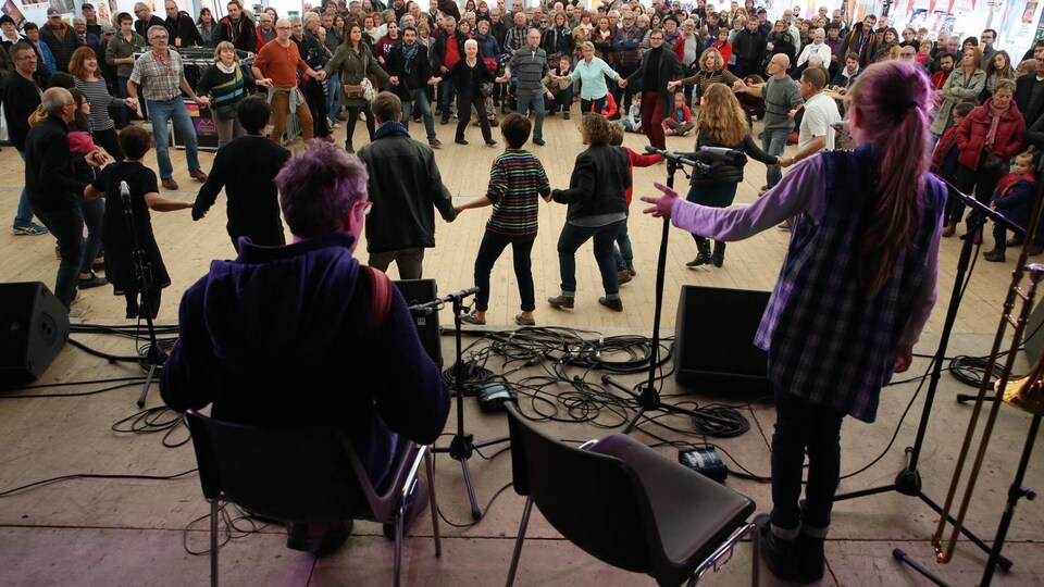 Un musicien et une jeune chanteuse sont sur scène devant un public qui se tiennent les mains en rond. 