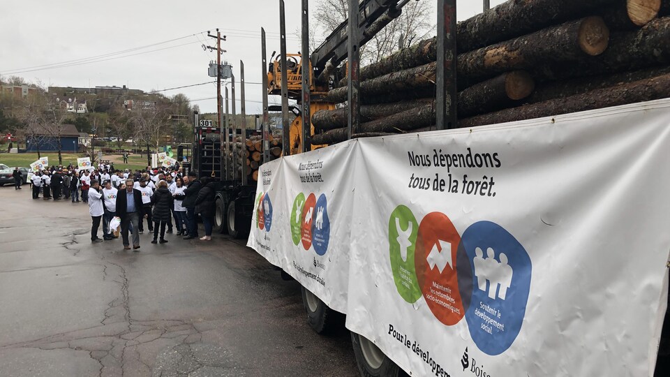 Un camion transporte des billots de bois et une affiche avec l'inscription « Nous dépendons tous de la forêt » est posée sur le côté. Des manifestants se trouvent en arrière-plan.