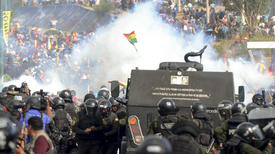 De la fumée s'élève entre des membres des forces de l'ordre portant un casque et des manifestants. Un drapeau de la Bolivie surgit au milieu de la fumée.