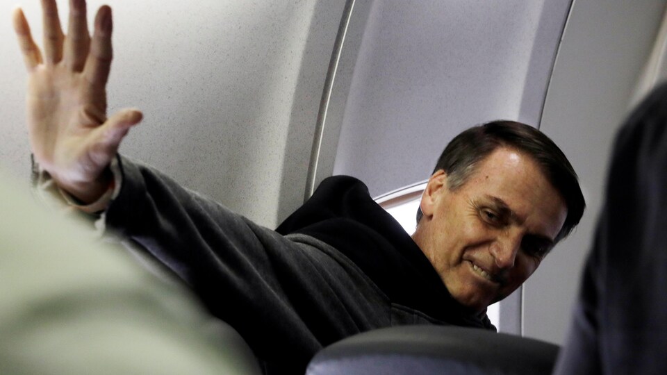 Jair Bolsonaro, assis dans l'avion, se tourne et salue de la main.