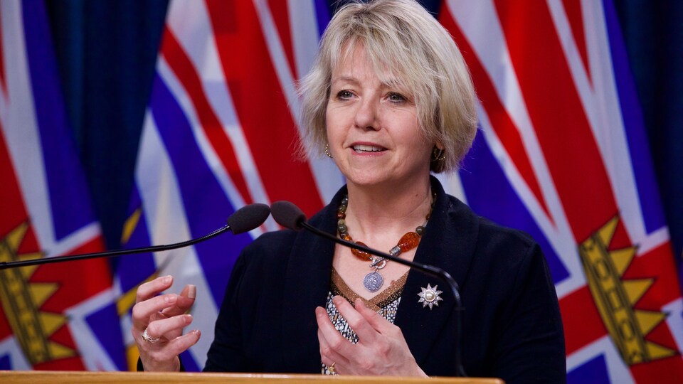 Bonnie Henry, en conférence de presse, devant des drapeaux de la Colombie-Britannique.