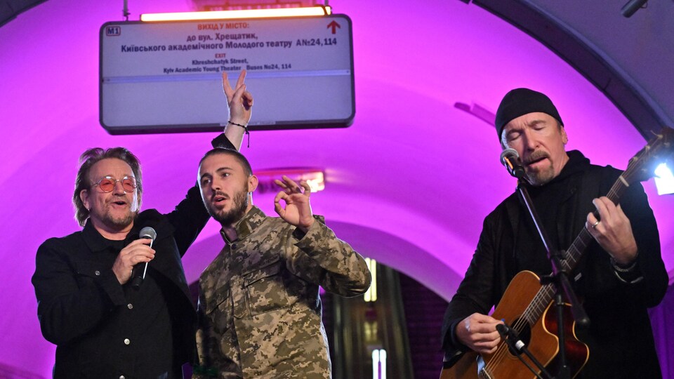 The three people, in a pink light, sing with a guitar and a microphone.
