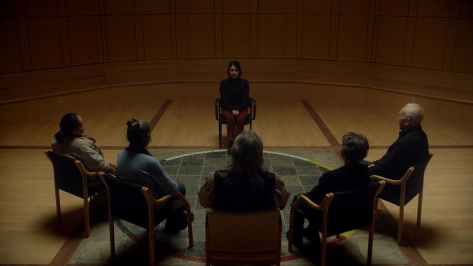 Une femme est assise devant cinq personnes assises en demi-cercle et vues de dos. 