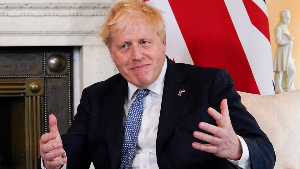 Boris Johnson, assis, les bras ouverts, la bouche fermée.