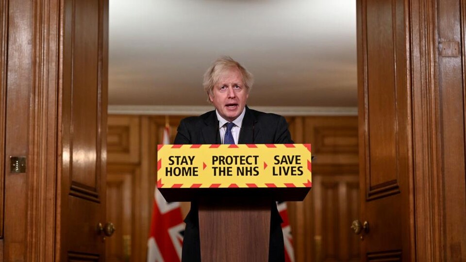 Boris Johnson se tient derrière un lutrin sur lequel on peut lire : « Restez à la maison, protégez le NHS [réseau de la santé], sauvez des vies. »
