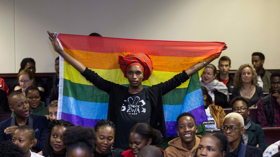Une personne debout dans une salle de cour tenant dans ses mains un drapeau arc-en-ciel.