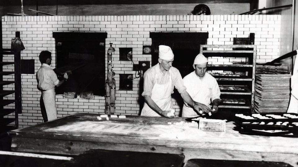 Photo en noir et blanc de boulangers qui manipulent de la pâte sur une grande table de travail avec une personne en train de mettre quelque chose au four en arrière.