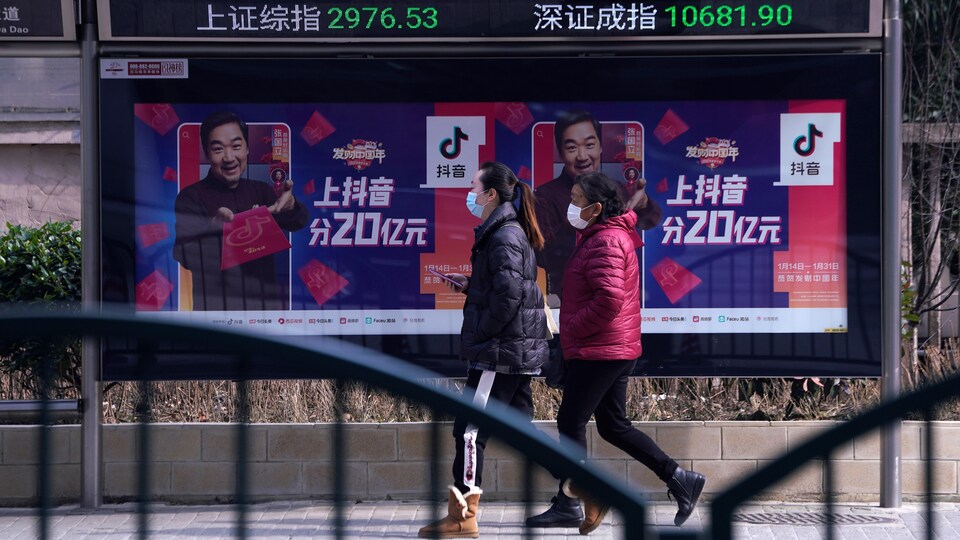 Deux femmes portant un masque marchent sur le trottoir devant un panneau d'affichage qui indique des données de la Bourse.