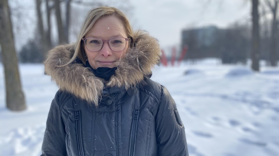 Une femme blonde qui porte un manteau d'hiver et des lunettes sourit à la caméra.