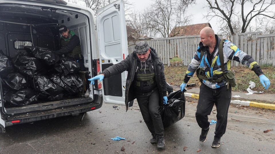 Deux hommes traînent un sac noir contenant un cadavre vers une fourgonnette chargée de dépouilles.