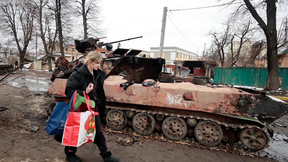 Une femme marche avec des sacs près d'un char militaire qui semble brûlé.