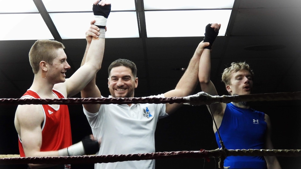 L'arbitre, entre deux boxeurs dans un ring, lève un bras de chacun d'eux en souriant.