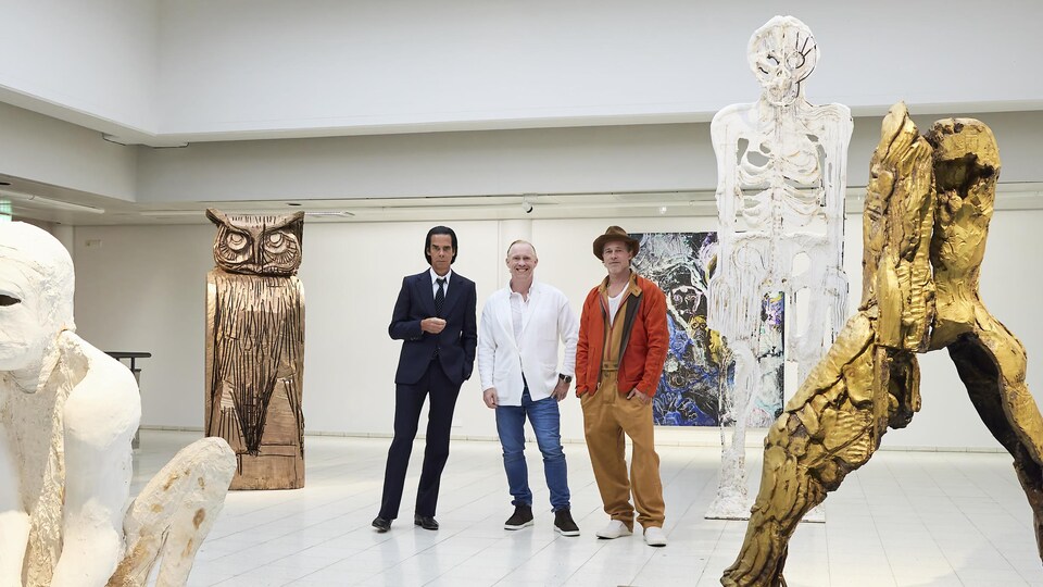 Trois hommes posent dans une galerie d'art jonchée de sculptures.