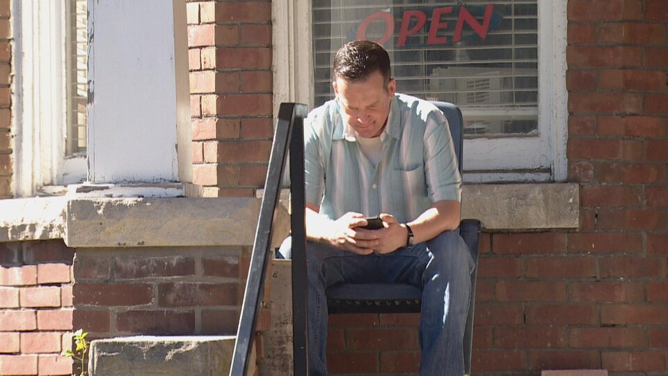Bradley Michael Finch, assis à l'extérieur devant un édifice, regardant son téléphone cellulaire.