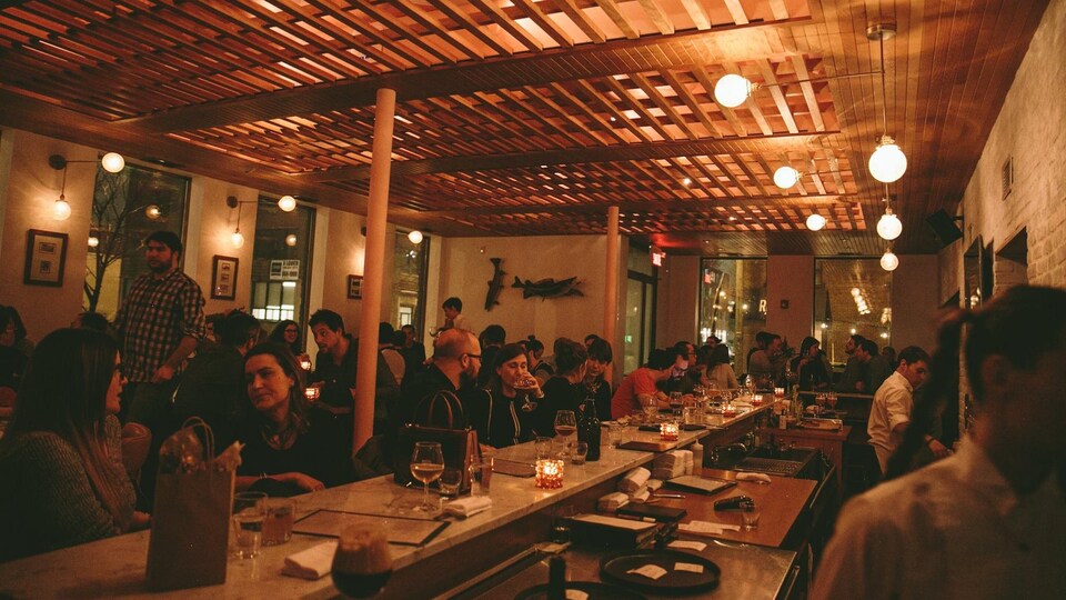 Des gens sont assis à des tables et au comptoir à l'intérieur d,une brasserie avec des lumières tamisées.