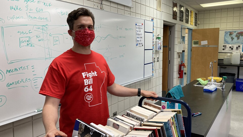 Un  homme portant un t-shirt rouge et un masque debout dans une salle de classe.