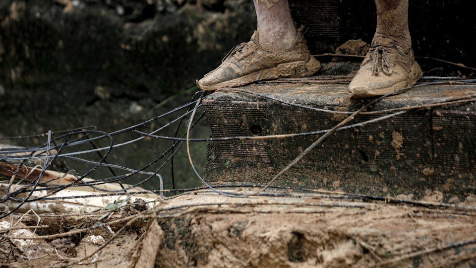 A man is sitting in front of a dilapidated house.  Mud covered his feet and ground.