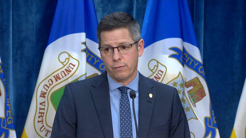 Brian Bowman lors de sa conférence de presse à l'hôtel de ville, devant un rideau bleu et des drapeaux de Winnipeg