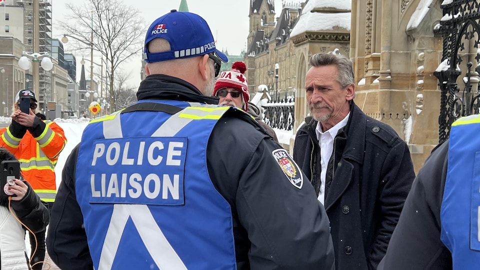 Brian Derksen est approché par un officier du Service de police d'Ottawa vêtu d'un dossard sur lequel il est écrit « Police Liaison », le 17 janvier 2023. Deux personnes autour d'eux filment la scène. 