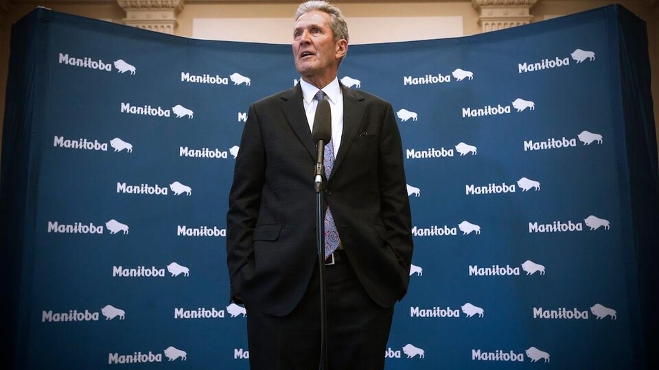 Brian Pallister parle, debout, derrière un micro et a les mains dans ses poches. On voit en arrière-plan plusieurs image du logo de la province (le mot Manitoba suivi d'une image stylisée de bison).