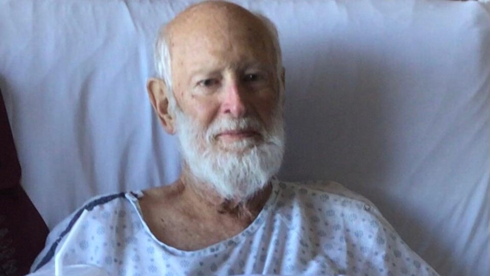 Un homme barbu est assis dans un lit d'hôpital. Il porte une chemise d'hôpital et tient un verre d'eau avec une paille.