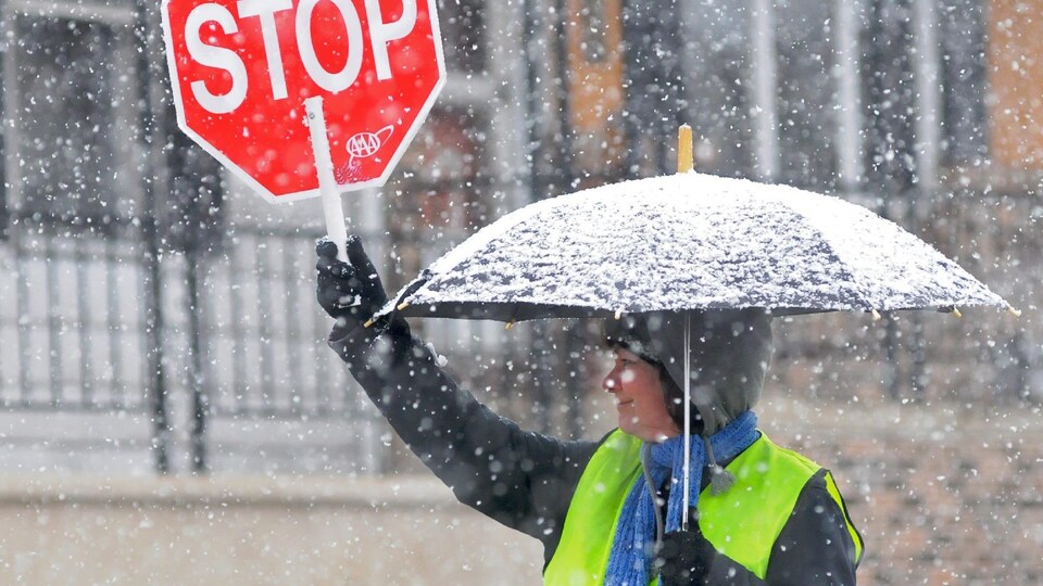 Une brigadière tient un panneau d'arrêt et un parapluie, tout sourire sous la neige.