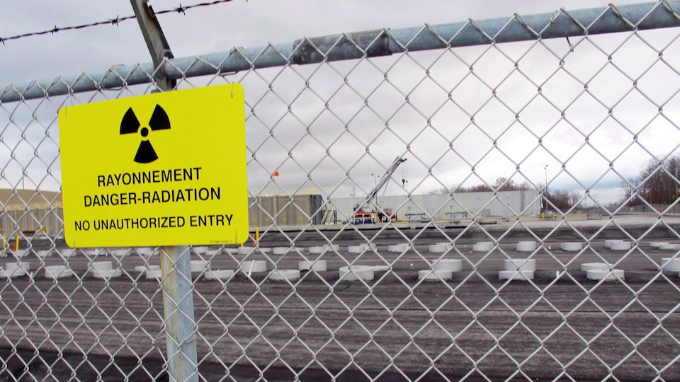 En avant-plan, une clôture avec une affiche où il est écrit : « Rayonnement Danger-Radioation, entrée non autorisée interdite ». En arrière-plan, une zone industrielle.