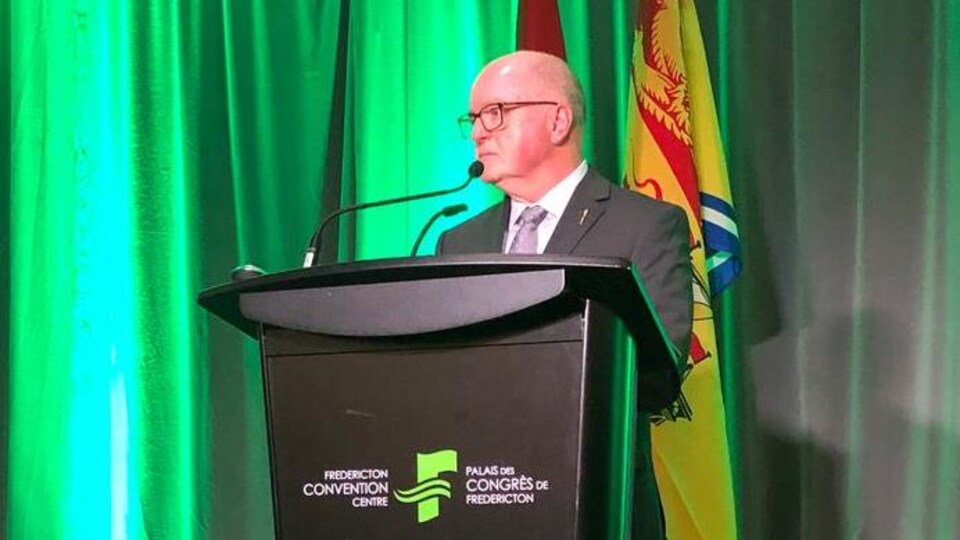 Bruce Fitch debout devant un lutrin du palais des congrès de Fredericton est baigné d'une vive lueur verte reflétée par les rideaux derrière lui et par le sommet de son crâne lisse et glabre.