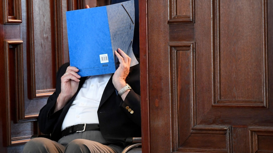 Un homme en chaise roulante se cache le visage avec un dossier bleu en franchissant une porte.