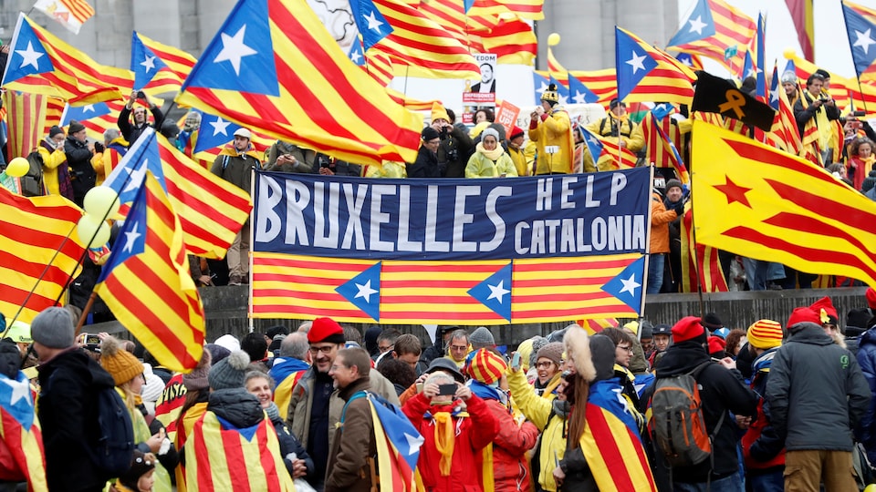 Des manifestants pro-Catalogne portent le drapeau catalan, rayé rouge et jaune, en guise de cape. Plusieurs autres drapeaux de la Catalogne flottent au vent.