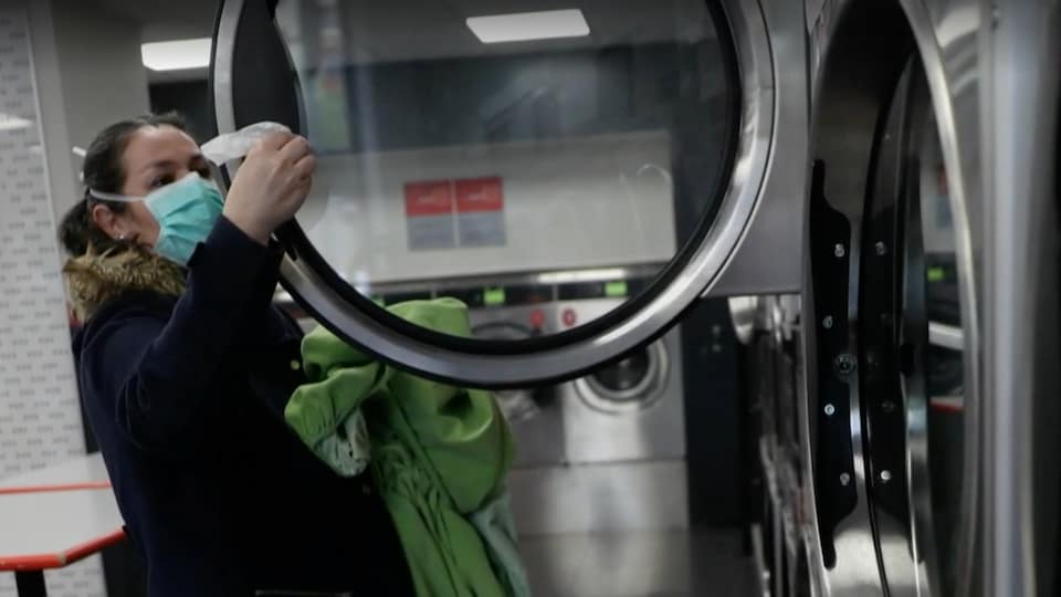 Une femme de profil portant un masque nettoie à l'aide d'une lingette l'intérieur d'une laveuse pour y déposes ses vêtements.