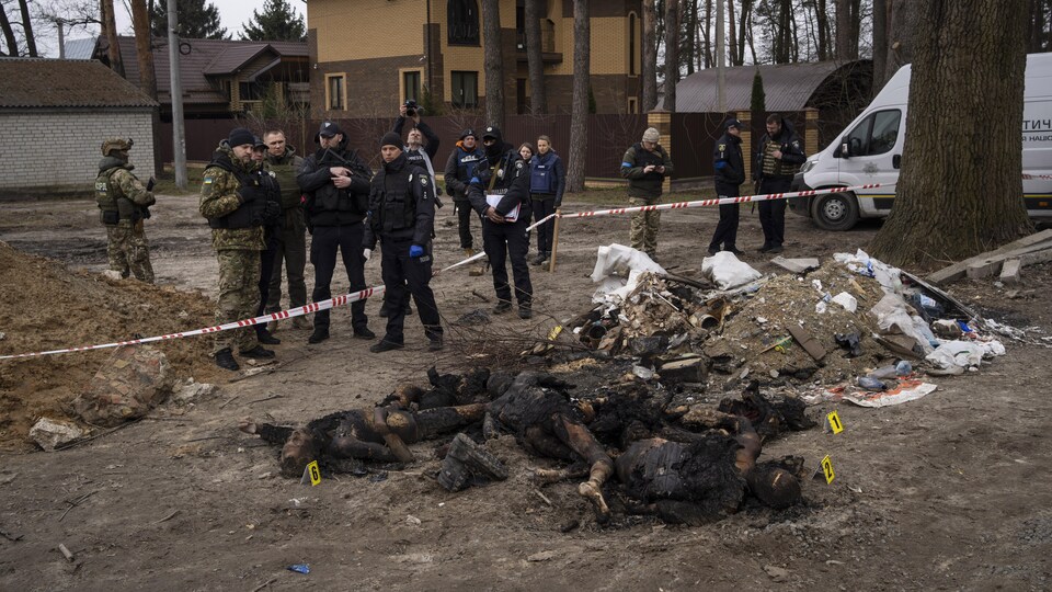 Des soldats et policiers se tiennent près d'un ruban qui les sépare de plusieurs corps entassés à demi brûlés.