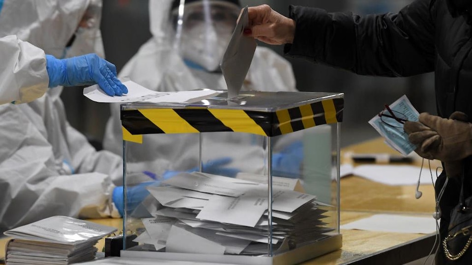 Des personnes vêtues de combinaisons intégrales, de masques, de visière et de lunettes de protection regardent une personne mettre son bulletin de vote dans l'urne.