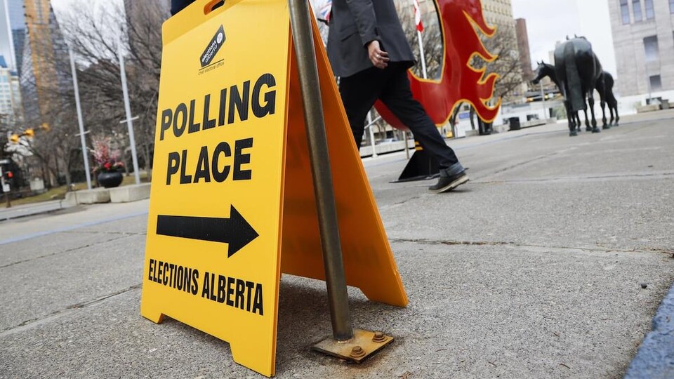 Un panneau indique un bureau de vote au centre-ville de Calgary pendant que des passants marchent sur un trottoir près d'un logo des Flames, l'équipe de hockey.