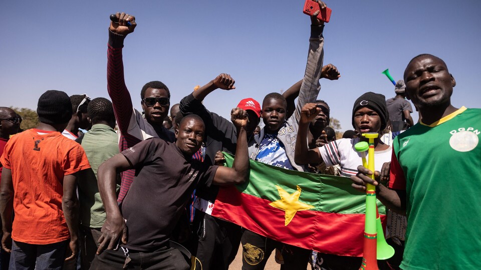 Des hommes, jeunes pour la plupart, célèbrent dans une rue. Ils tiennent un drapeau du Burkina Faso et ont le poing levé en signe de victoire. 