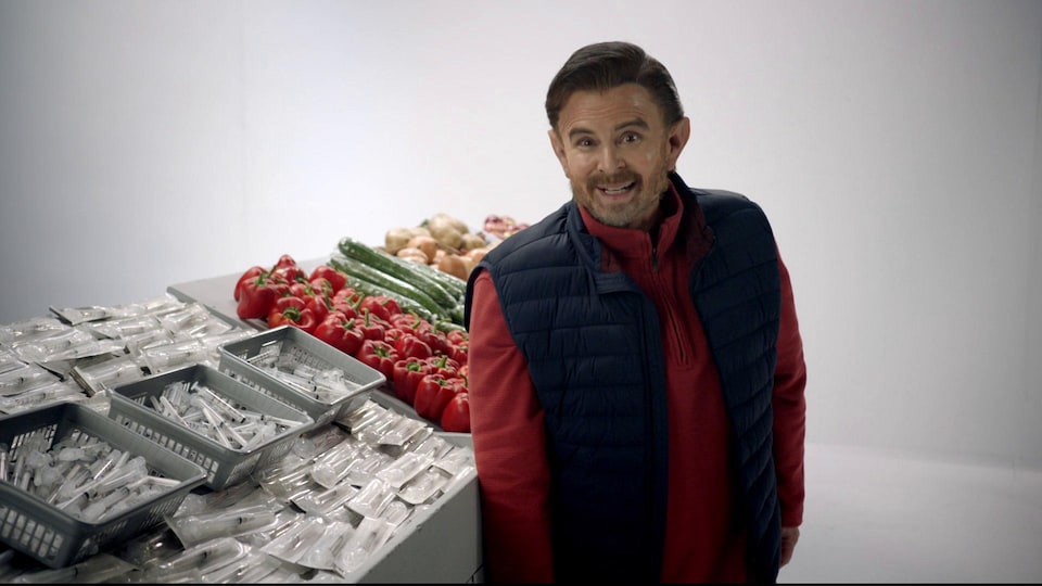 Un homme se tient devant un étal de marché rempli de seringues et de légumes.