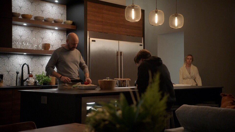Le comédien et deux comédiennes se tiennent debout dans une cuisine moderne. L'homme coupe des aliments.