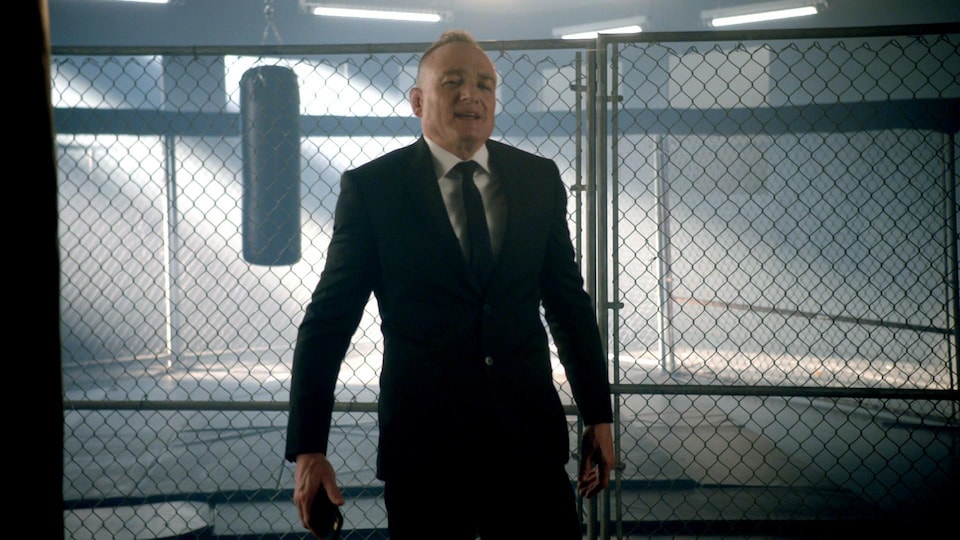 Un homme en complet cravate se tient devant une clôture grillagée dans une salle de boxe.