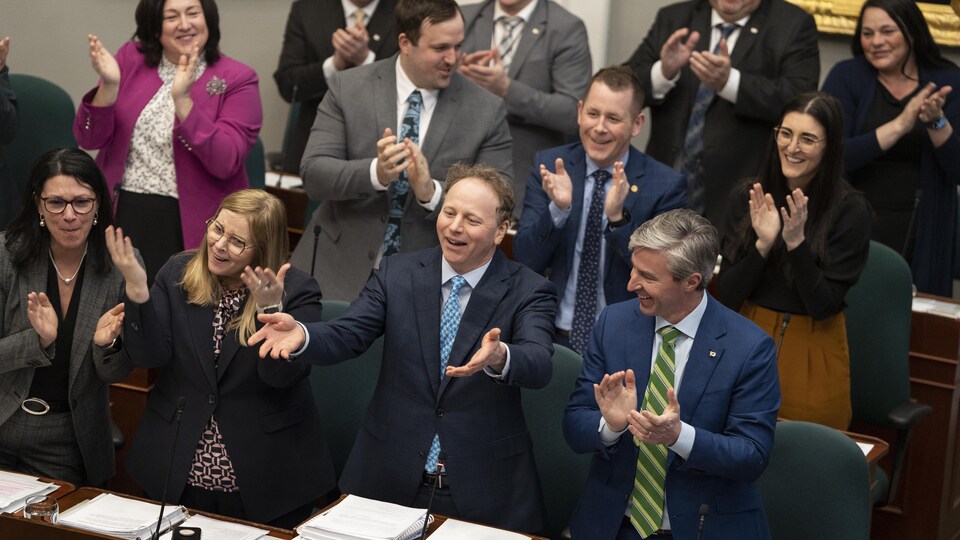 Le premier ministre et des membres de son Cabinet sont debout à l'Assemblée législative. Ils applaudissent le ministre des Finances, qui est au milieu, les deux bras tendus devant lui et la bouche ouverte.