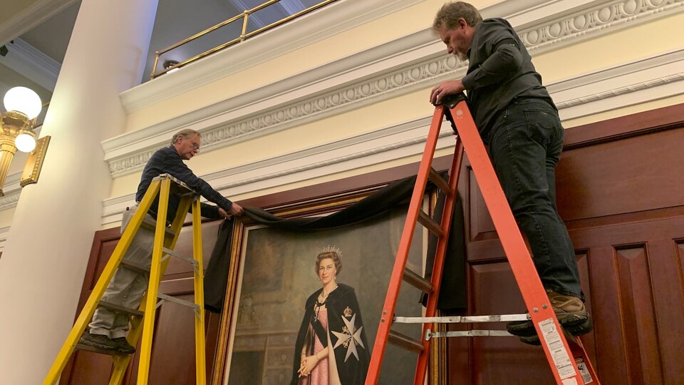 Deux hommes sur des échelles en train de mettre un drap noir sur le portrait de la reine Élisabeth II.
