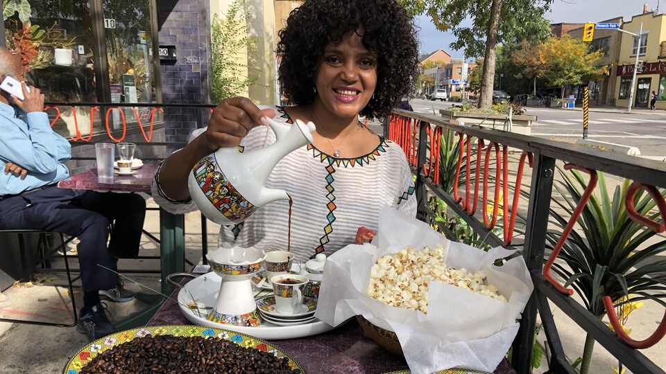 Une femme assise à la terrasse devant un homme qui parle au téléphone. Jebena à la main, elle verse le café dans des petites tasses rangées sur un plateau à coté de grains de café et de maïs soufflé.