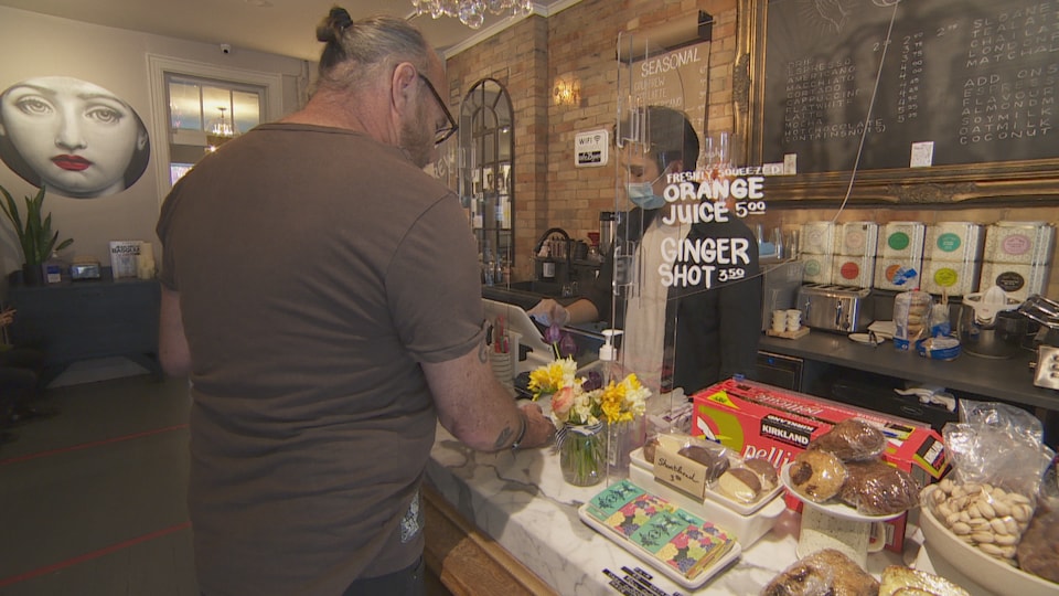 Un homme devant un barista, séparés par un écran en plexiglas, dans un café.