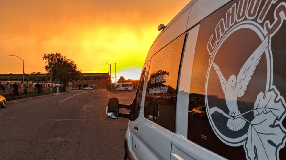 Une camionnette blanche aux vitres noires, avec un grand logo "CAHOOTS" et un oiseau blanc. Au fond, un coucher de soleil.
