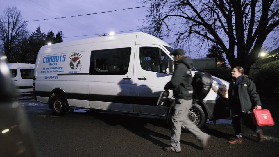 Une camionnette blanche au logo "CAHOOTS", le soir. À droite, deux personnes transportent des équipements.