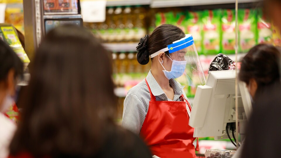 Une caissière de supermarché portant un masque, une visière et des gants.