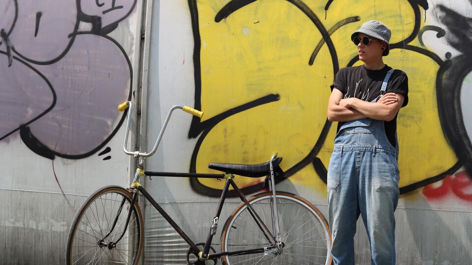 Une femme croise les bras à côté de son vélo, posé contre un mur rempli de graffitis. 