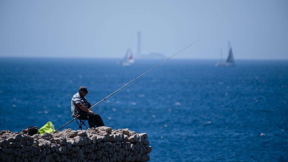 Un homme est assis au bord d'une calanque, où il pêche.