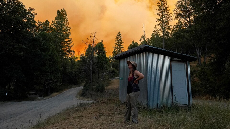 Une femme se tient debout devant une cabane alors qu'un incendie de forêt enfume le paysage.